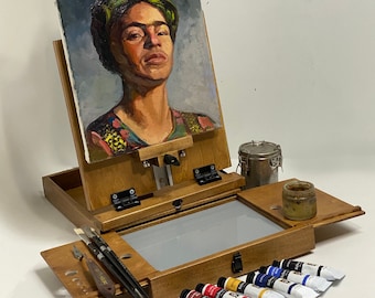 May include: A wooden tabletop easel holding an oil painting of a person with a green headpiece. The easel features a fold-out tray containing three paintbrushes, a palette knife, a small jar, and six tubes of oil paint labelled Richeson.