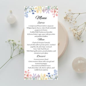 May include: A rectangular menu card with floral decorations at the top and bottom. The menu lists Entrée, Main, and Dessert courses with detailed descriptions. The card is placed on a light pink plate.