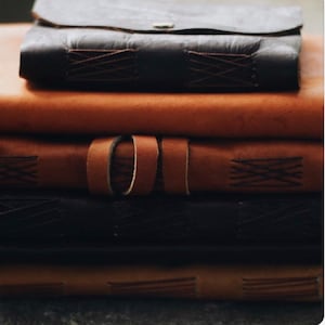 May include: A stack of five leather-bound journals with brown and black covers. The journals have decorative stitching and are arranged in a pile.