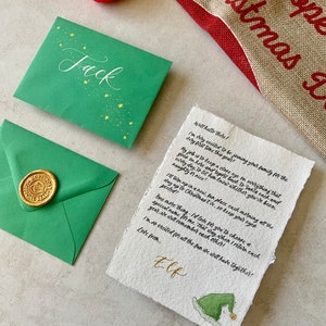 May include: A handwritten letter addressed to Jack from an elf. The letter is written on a piece of parchment paper with a green elf hat illustration at the bottom. The letter mentions keeping a close eye on everything that goes on and reporting back to Santa each day.