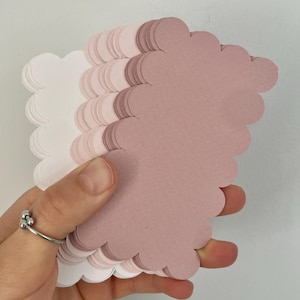 Op de afbeelding: Een stapel wolk-vormige papieren uitsparingen in verschillende tinten roze en wit. Het papier heeft een geschulpte rand. De uitsparingen worden in een hand gehouden tegen een effen witte achtergrond.