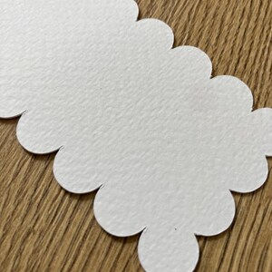 Op de afbeelding: Een witte papieren tag met geschulpte randen en een getextureerd oppervlak. De tag is uitgesneden in een lange, onregelmatige vorm met afgeronde randen. De achtergrond is een houten oppervlak.