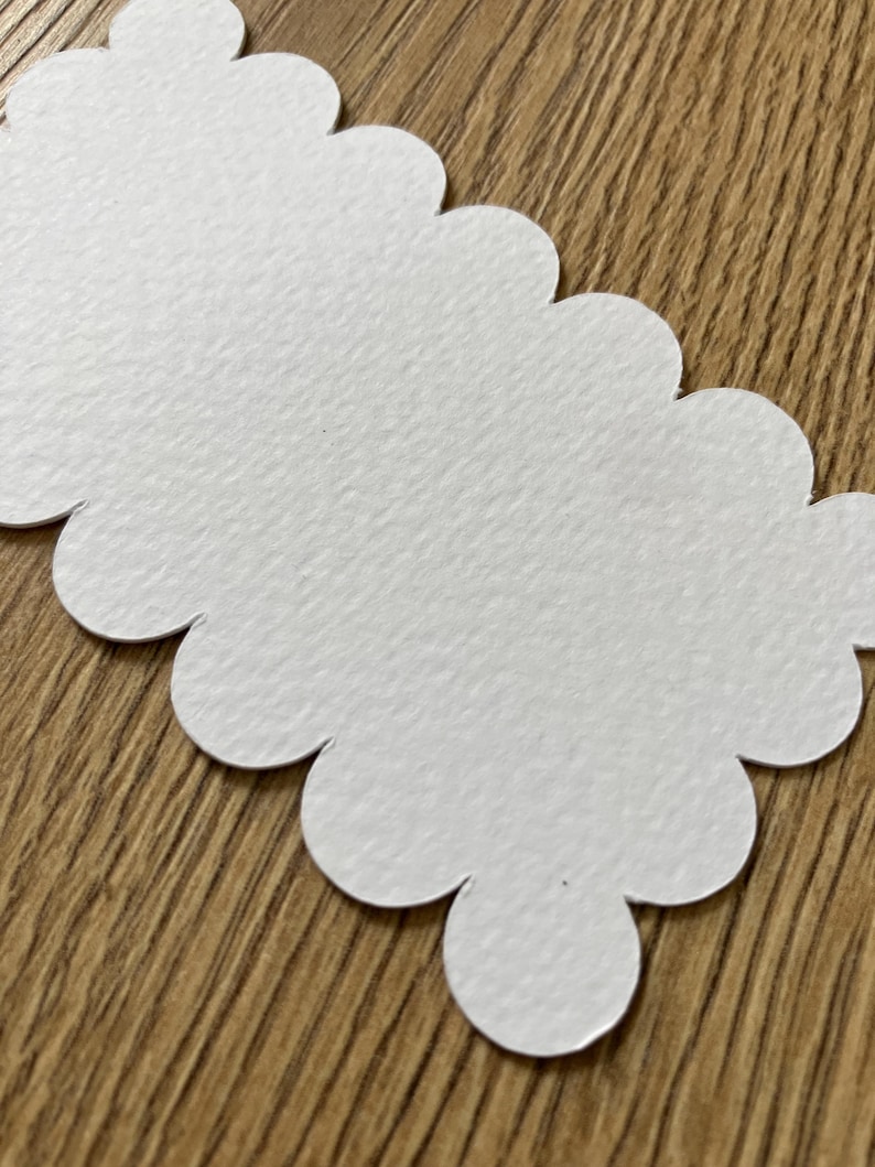 Op de afbeelding: Een witte papieren tag met geschulpte randen en een getextureerd oppervlak. De tag is uitgesneden in een lange, onregelmatige vorm met afgeronde randen. De achtergrond is een houten oppervlak.