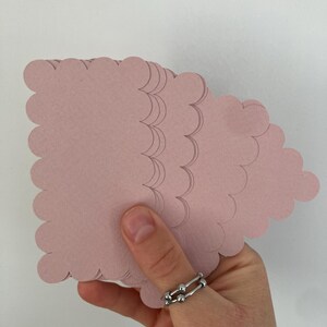 Op de afbeelding: Een hand die een stapel roze papieren uitsparingen in de vorm van een wolk vasthoudt. Het papier is zachtroze van kleur en heeft een geschulpte rand. Er wordt een zilveren ring aan de hand gedragen.