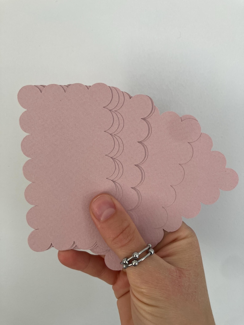 Op de afbeelding: Een hand die een stapel roze papieren uitsparingen in de vorm van een wolk vasthoudt. Het papier is zachtroze van kleur en heeft een geschulpte rand. Er wordt een zilveren ring aan de hand gedragen.