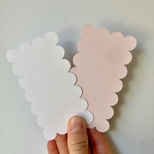 Op de afbeelding: Een hand houdt twee wolk-vormige papieren uitsparingen vast, &eacute;&eacute;n witte en &eacute;&eacute;n lichtroze. De uitsparingen hebben een geschulpte rand. De achtergrond is een effen wit oppervlak.