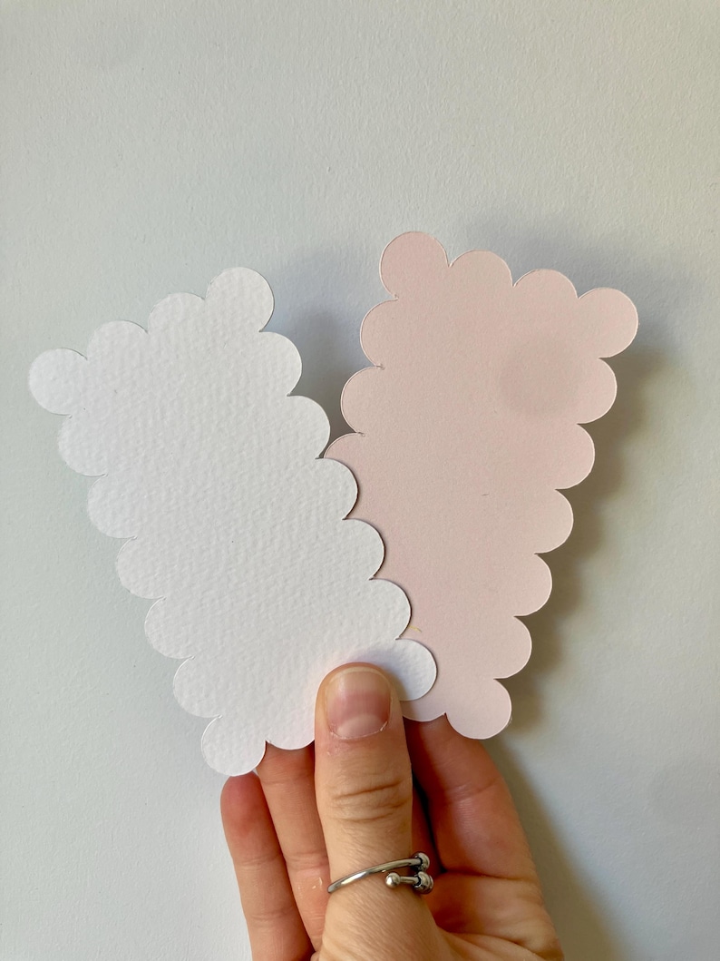 Op de afbeelding: Een hand houdt twee wolk-vormige papieren uitsparingen vast, &eacute;&eacute;n witte en &eacute;&eacute;n lichtroze. De uitsparingen hebben een geschulpte rand. De achtergrond is een effen wit oppervlak.