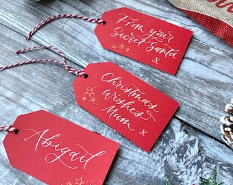 May include: Three red gift tags with white calligraphy. The top tag says "From your Secret Santa." The middle tag says "Christmas Wishes Mum." The bottom tag says "Abigail." All three tags have gold stars and a white string.