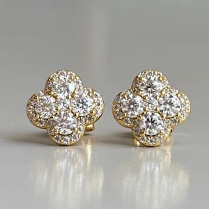 Puede incluir: Un par de pendientes dorados, cada uno con un diseño de trébol de cuatro hojas. Los pendientes están incrustados con múltiples gemas claras y brillantes, con un halo de piedras más pequeñas alrededor de las más grandes.