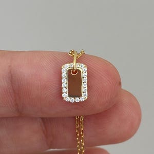 May include: A gold-coloured rectangular pendant necklace with a delicate chain. The pendant is outlined with small, clear gemstones. The necklace is held against a neutral background, showcasing the jewellery's design and details.