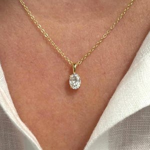 May include: A delicate gold chain necklace featuring a single, small, oval-cut clear gemstone pendant. The pendant is set in a simple gold prong setting, suspended from a thin, polished gold cable chain that rests against a white collared shirt.