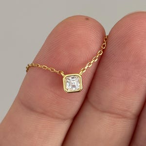 May include: A delicate gold necklace featuring a square, clear gemstone pendant. The pendant is set in a gold bezel and hangs from a fine gold chain. The necklace is shown against a neutral background.