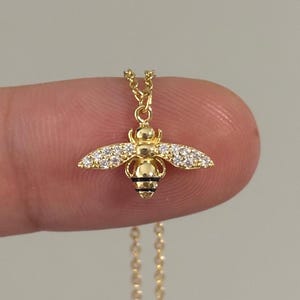 May include: A gold-coloured necklace with a bee pendant. The bee has crystal-like detailing on its wings and a black and gold body. The necklace chain is fine and gold, creating a simple and elegant design.