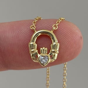 May include: A gold Claddagh necklace. The pendant shows a heart, hands, and a crown. The necklace is held by a finger against a plain background. The chain is fine and gold-coloured.
