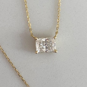 Könnte beinhalten: Eine zarte goldene Halskette mit einem rechteckigen, klaren Edelsteinanhänger. Der Anhänger ist in einer goldenen Fassung gefasst und hängt an einer feinen Goldkette. Die Halskette wird auf einer hellen Oberfläche präsentiert.