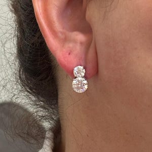 May include: A pair of silver-toned stud earrings, each featuring two vertically aligned round, clear, faceted stones. The earrings are secured to the earlobe with a post-back mechanism, showcasing a bright, reflective, and polished metallic setting.