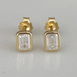 May include: A pair of gold stud earrings with rectangular, clear, gemstone centers. The earrings are set in a gold bezel setting. The earrings are displayed on a white and gray marble surface.