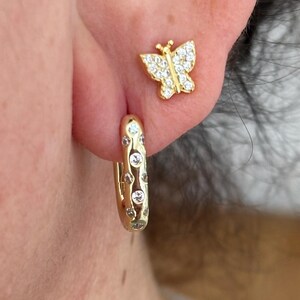 May include: A person's ear is shown, displaying two pieces of jewellery. A gold butterfly stud earring sits above a gold hoop earring. The hoop earring has small, clear stones. A gold chain necklace is also visible.