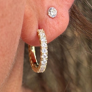 May include: Close-up of a person's ear adorned with two pieces of jewelry. A small, round, clear gemstone stud earring is in the upper ear. A gold-colored hoop earring with multiple clear gemstones is in the lower ear.