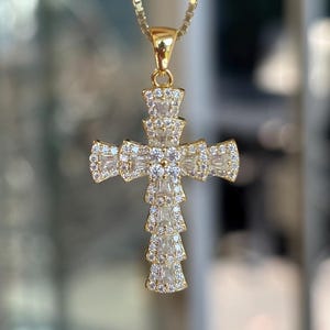 May include: A gold-coloured cross pendant necklace with a fine chain. The cross is adorned with clear, sparkling gemstones. The pendant is a religious symbol, suitable for everyday wear or special occasions.