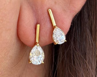 May include: A close-up of a person's ear adorned with two gold earrings. Each earring features a small, vertical gold bar connected to a pear-shaped, clear gemstone. The earrings are set against the backdrop of the person's skin and hair.