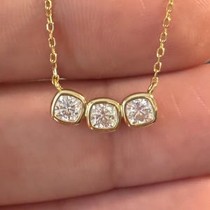 May include: A gold necklace with three square, clear gemstones. The gemstones are set in individual bezels and are connected by a fine gold chain. The necklace is displayed against a blurred background.