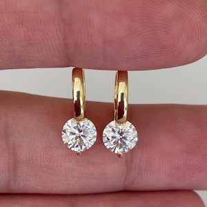 May include: Two gold-coloured hoop earrings, each with a large, round, clear gemstone. The earrings are displayed against a plain background, highlighting their simple, elegant design. The gemstones sparkle, reflecting the light.