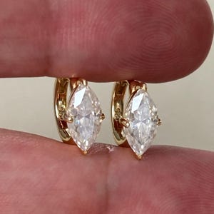 Puede incluir: Un par de pendientes de aro dorados, cada uno con una piedra preciosa transparente de talla marquesa engastada en garras. Los pendientes son aros circulares pequeños con un acabado en oro pulido y una piedra tallada alargada.