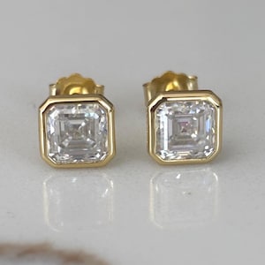 May include: A pair of gold-colored square stud earrings with a clear, square-cut gemstone in the center. The earrings are set in a gold bezel setting and are displayed on a white surface.