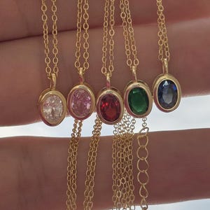 May include: Six gold necklaces, each with an oval gemstone in clear, pink, red, green, and blue. The gemstones are set in gold bezels and suspended from fine gold chains. The necklaces are shown against a blurred backdrop.