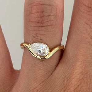 May include: A gold ring featuring a single pear-shaped white gemstone set in a smooth, curved band. The metal band has a polished finish and follows a wave-like design that cradles the faceted stone, creating a sleek and continuous appearance.