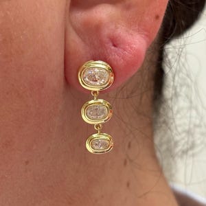 May include: A gold drop earring featuring three vertically aligned oval-shaped clear stones, each encased in a smooth gold bezel setting. The earring hangs from the earlobe, showcasing a polished metallic finish and transparent faceted gems.