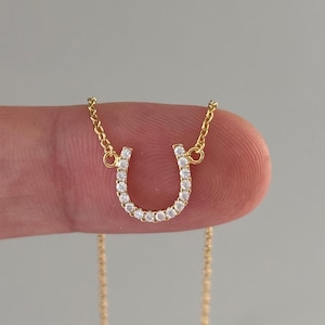 May include: A gold-colored horseshoe pendant necklace. The horseshoe is encrusted with small, clear gemstones. The necklace chain is also gold-colored and delicate. The pendant is held up by a finger against a gray background.