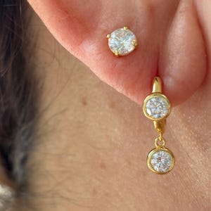May include: A gold stud earring featuring a single round clear stone and a gold hoop earring with two dangling round clear stones. The jewellery is crafted with a polished gold finish and secure settings for each of the sparkling white stones.