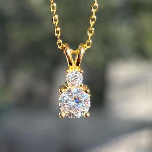 May include: A gold necklace with a pendant. The pendant has a large, round, clear gemstone and a smaller gemstone above it. The chain and pendant setting are gold. The background is blurred.
