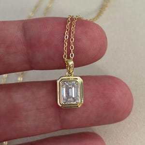 May include: A gold-tone necklace featuring a rectangular emerald-cut clear stone pendant set in a bezel frame. The pendant hangs from a delicate gold-tone chain. The text RaffEssentials is visible in the lower left corner of the image.