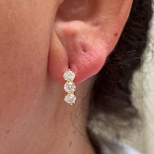 May include: A gold hoop earring featuring three round, clear, faceted stones set in a vertical row. The earring is secured through the earlobe, showcasing a polished gold metal finish and brilliant, reflective white stones in a classic design.