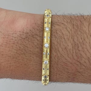 Op de afbeelding: Een goudkleurige armband met een getextureerd oppervlak en decoratieve elementen. De armband heeft een herhalend patroon van kleine, heldere edelstenen en bloemmotieven. De armband is ongeveer 1,3 cm breed.