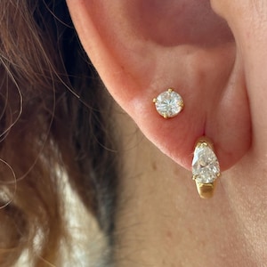 May include: A gold stud earring featuring a round brilliant-cut clear stone and a gold hoop earring featuring a pear-shaped clear stone. Both earrings are crafted from yellow gold and are positioned in the earlobe, showcasing a classic design.