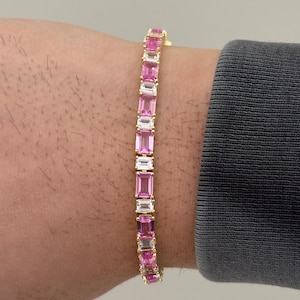 May include: A gold bracelet featuring alternating pink and clear rectangular gemstones. The bracelet is worn on a wrist, showcasing the design and color contrast. The gemstones are set in a gold band.