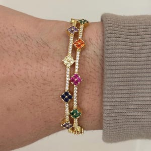 Puede incluir: Pulsera dorada con una serie de gemas de colores en forma de flor. La pulsera tiene un diseño delicado con acentos claros y brillantes entre las gemas. Las gemas son en tonos verdes, azules, rosas, naranjas y morados.