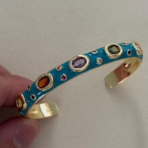 May include: A gold-tone cuff bracelet featuring a bright turquoise enamel finish. The surface is inlaid with various multi-coloured gemstones in oval and round shapes, including purple, orange, yellow, and clear stones set in gold bezels.