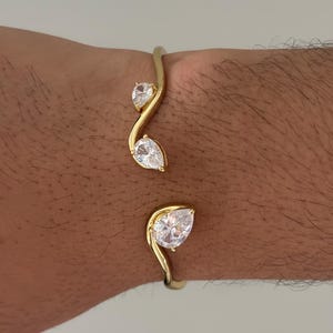 Puede incluir: Una pulsera de brazalete abierto en tono dorado con tres piedras de circonita cúbica transparentes en forma de pera. La banda tiene un diseño curvo y ondulado, con dos piedras en un extremo y una piedra más grande en el extremo opuesto.