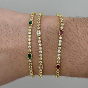 Peut inclure: Trois bracelets dorés ornés d'une rangée de petites pierres transparentes. Chaque bracelet comporte une pierre précieuse ovale plus grande : une verte, une transparente et une rouge. Les bracelets sont présentés sur le bras d'une personne.