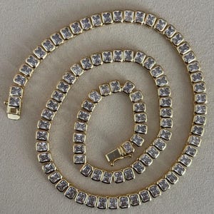 May include: A gold-toned necklace featuring a series of square-cut, clear gemstones. The necklace is laid out in a spiral, showcasing the clasp and the intricate setting of each stone. The overall design is elegant and suitable for special occasions.