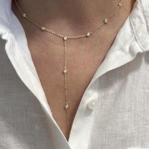May include: A gold Y-shaped lariat necklace featuring a delicate chain adorned with multiple evenly spaced round bezel-set clear stones. The necklace rests against the collarbone and extends downward in a vertical drop, creating a minimalist aesthetic.