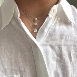 May include: Three diamond necklaces of varying sizes are layered on a person's neck. The diamonds are round-cut and set in silver or white gold. The person is wearing a white button-down shirt.