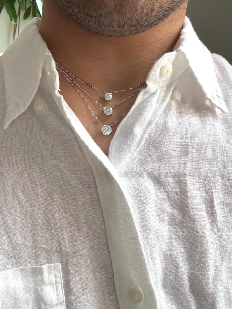 May include: Three diamond necklaces of varying sizes are layered on a person's neck. The diamonds are round-cut and set in silver or white gold. The person is wearing a white button-down shirt.