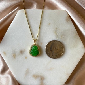 May include: A gold chain necklace with a green jade Buddha pendant. The pendant is surrounded by small clear stones.
