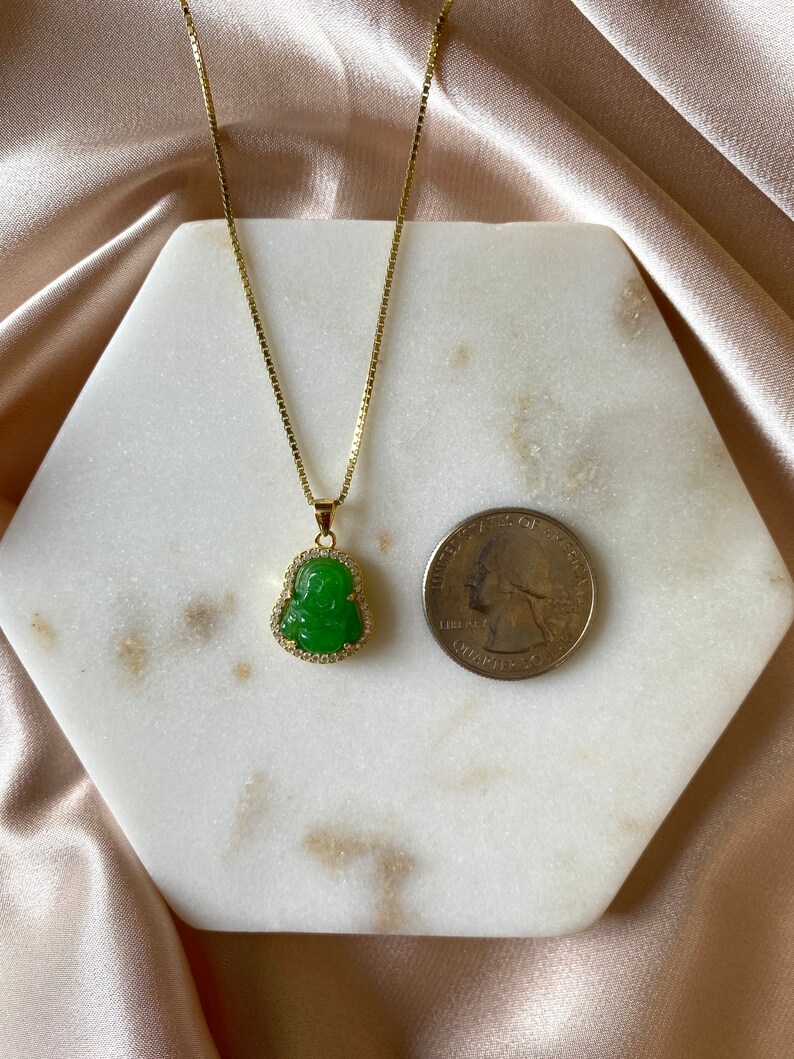 May include: A gold chain necklace with a green jade Buddha pendant. The pendant is surrounded by small clear stones.